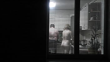 Peeping. Voyeur. Neighbor freak Spycam in evening on street looks out window as nude super-sexy neighbor in kitchen preps dinner for her husband. nude in public. nude at home. Family.  Outdoor